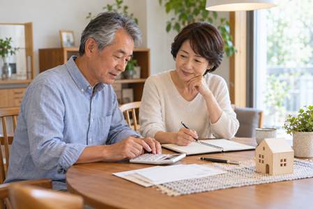 夫婦がテーブルに置いた電卓とノートを使い、住宅模型を前に家計と住まいの予算を相談する様子。